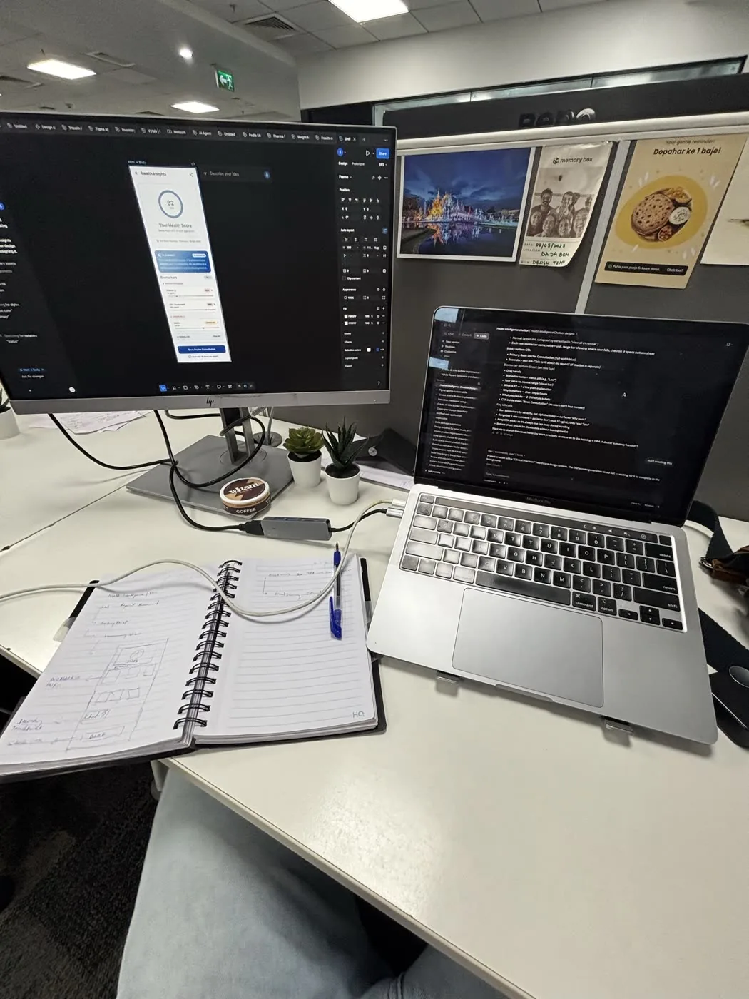 A desk: a monitor running Figma, a laptop beside it, and an open notebook of hand-drawn wireframes with a pen across the page.